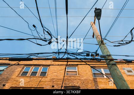 I cavi elettrici si allungano nel cielo sopra un edificio in mattoni. Un palo alto si trova di lato. Il cielo blu aggiunge contrasto alla scena durante il giorno. Foto Stock