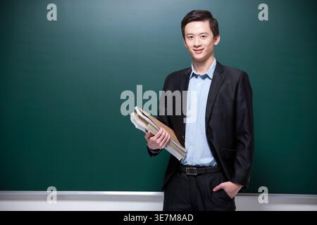 Un insegnante cinese sorridente che indossa una tuta nera che tiene in mano un libro mentre si sta in piedi con la mano in tasca contro la lavagna in classe Foto Stock