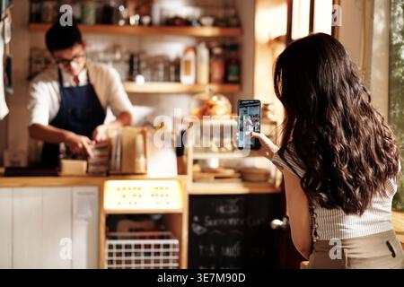 Donna asiatica con lunghi capelli ricci che fotografa un barista che lavora al bancone del bar Foto Stock