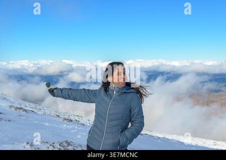 Donna ispanica di mezza età sorridente in giacca calda con occhiali da sole e sguardo lontano mentre si sta in piedi con la mano in tasca sulla montagna innevata con... Foto Stock