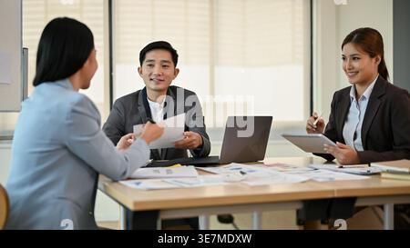 Uomo d'affari asiatico sorridente che lavora insieme a colleghi seduti a tavola e che discute relazioni finanziarie in ufficio Foto Stock