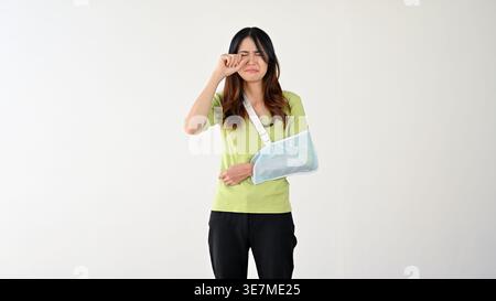 Triste donna asiatica che si pulisce la lacrima indossando un'imbracatura per il braccio in piedi su sfondo grigio Foto Stock