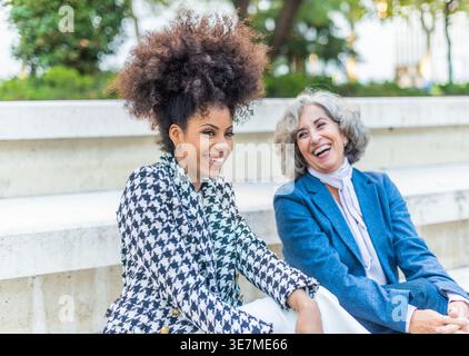 Donna ispanica con acconciatura afro che ride con anziana donna caucasica seduta su una panchina di cemento nel parco durante il giorno Foto Stock