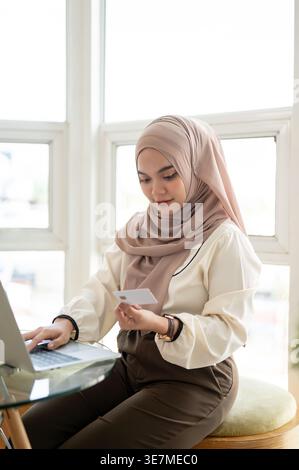 Donna musulmana sorridente che indossa hijab usando la sua carta di credito per acquistare online usando il laptop mentre si siede a un tavolo di vetro vicino alla finestra di casa Foto Stock