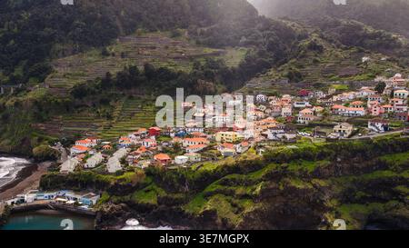 Vista aerea di un villaggio dell'isola di Madeira con case bianche e tetti arancioni su ripide scogliere sopra un piccolo porto e spiaggia di sabbia nera, campi terrazzati, Foto Stock
