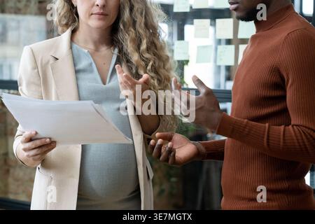 Uomo d'affari nero in piedi accanto a una collega bianca incinta che tiene i documenti in carica. Foto Stock