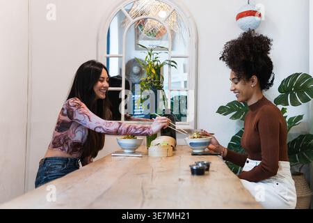 Amiche ispaniche e miste che si divertono a mangiare e a chiacchierare mentre si siedono al tavolo di legno nel ristorante Foto Stock