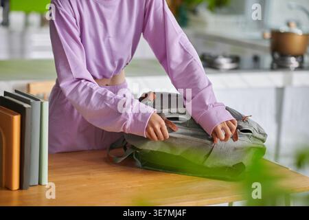 Donna asiatica che indossa uno zaino con cerniera superiore a maniche lunghe viola che si prepara per la scuola mentre è in piedi a tavola in cucina a casa Foto Stock
