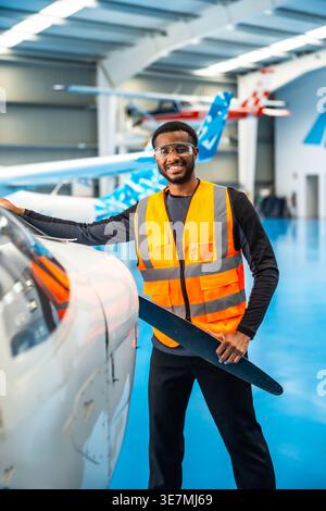 Sorridente ingegnere afroamericano maschio di aerei che tiene la lama dell'elica nell'hangar Foto Stock