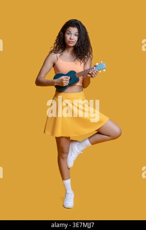 Donna afroamericana che gioca a ukulele in piedi con un piede su sfondo giallo Foto Stock