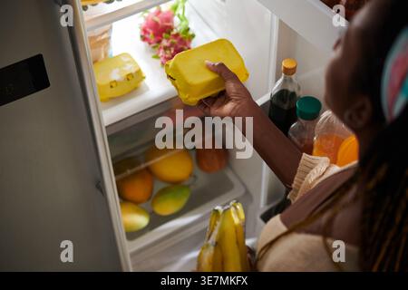 Donna nera che prende una scatola di uova dal frigorifero in cucina a casa Foto Stock