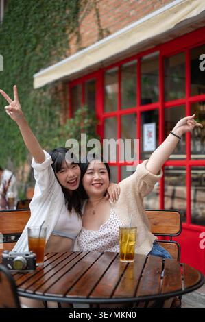 Allegre amiche asiatiche che alzano la mano con un cartello di pace che beve sedute al tavolo in un bar Foto Stock