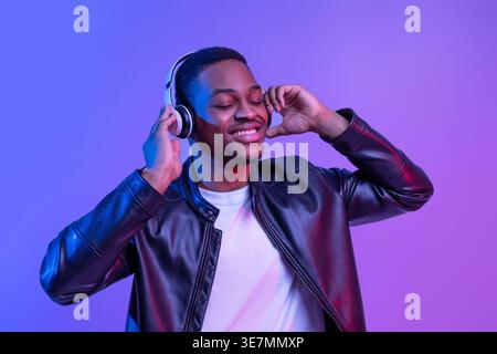 Uomo nero sorridente che indossa una giacca in pelle che ascolta la musica tramite le cuffie wireless mentre sei in piedi con gli occhi chiusi contro lo schienale viola al neon... Foto Stock