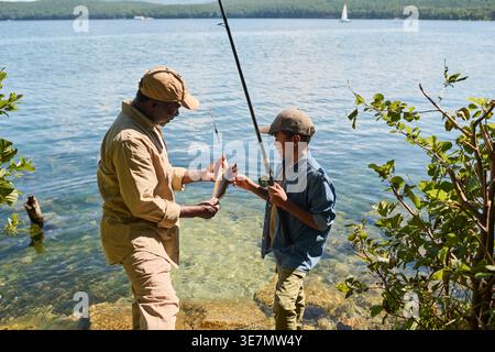 Vista laterale dell'uomo nero di mezza età e del nipote felice che tiene la canna e il pesce appena pescato in piedi sul bordo del lago durante il campeggio nel... Foto Stock