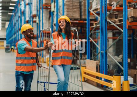 Lavoratore nero che indossa elmetto e giubbotto di sicurezza divertendosi a spingere il metallo cLifestyle con una collega caucasica in piedi su di esso in magazzino Foto Stock