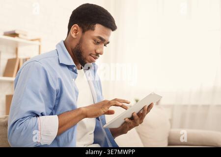 Uomo nero che usa un tablet seduto sul divano di casa Foto Stock