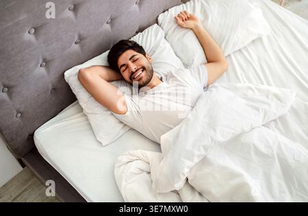 Uomo arabo sorridente con la barba che indossa una camicia bianca che allunga le braccia con gli occhi chiusi mentre è sdraiato sul letto in camera da letto di casa Foto Stock