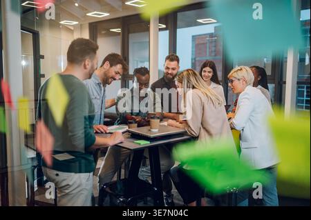 Uomo d'affari di colore che tiene in mano la festa del telefono con i colleghi intorno al tavolo in ufficio Foto Stock