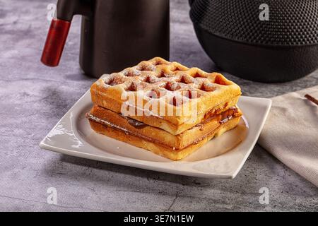 Una pila di waffle appena sfornati con ripieno di cioccolato e zucchero a velo su un piatto bianco. Colazione accogliente o brunch in uno sfondo grigio Foto Stock
