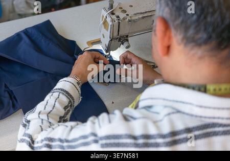 Vista posteriore del sarto ispanico di mezza età che cucisce un orlo di pantaloni sulla macchina da cucire mentre si siede in un negozio di sartoria Foto Stock