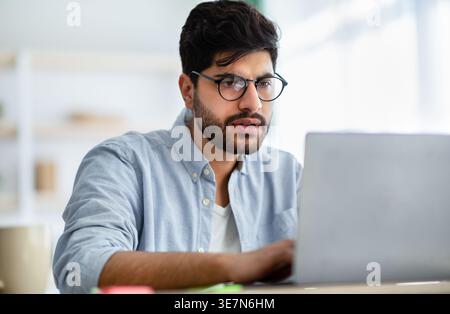 Uomo arabo con la barba che indossa gli occhiali che lavora su un computer portatile seduto alla scrivania di casa Foto Stock