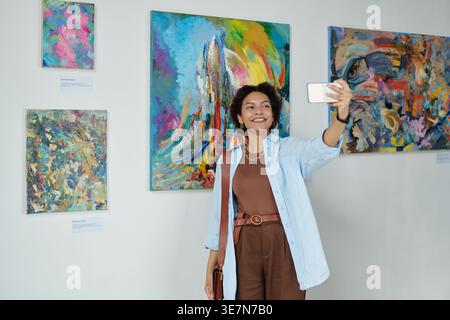 Adolescente nera che scatta selfie contro i dipinti mentre è in piedi nella galleria d'arte Foto Stock
