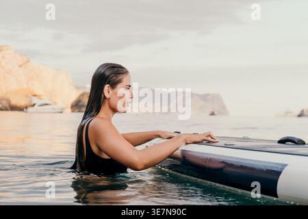 Donna Paddleboarding Ocean - Una donna con un costume da bagno nero fa paddleboarding nell'oceano. Sta guardando di lato, e l'acqua è calma. Qui Foto Stock