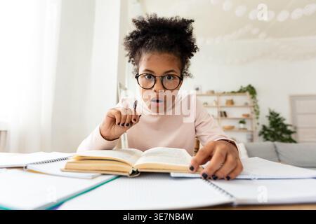 Scioccata ragazza nera preadolescente che indossa gli occhiali tenendo la penna seduta a tavola con un libro aperto a casa Foto Stock