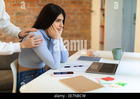 Triste studentessa mediorientale con la mano della madre sulla spalla seduta alla scrivania con il portatile e i materiali di studio nel soggiorno di casa Foto Stock