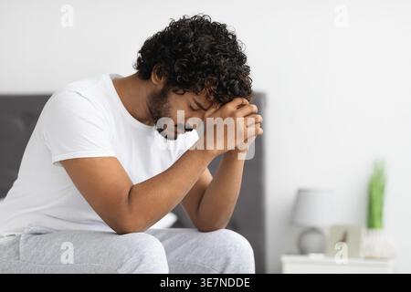Uomo indiano barbuto stressato con capelli ricci appoggiati sulla fronte su pugni clenati seduti sul letto in camera da letto di casa Foto Stock