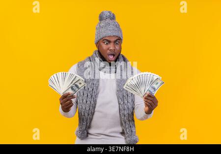 Scioccato uomo Nero che indossa berretto e sciarpa a maglia che guarda i soldi nelle sue mani in piedi su sfondo giallo in studio Foto Stock