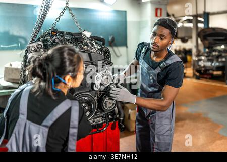 Meccanici neri maschi e asiatici che indossano uniformi e occhiali protettivi che ispezionano il motore di un'auto appeso a un paranco a catena in un negozio di riparazione auto Foto Stock