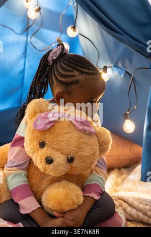 Ragazza afroamericana in età scolare che abbraccia un orsacchiotto con fiocco rosa in tenda blu, luci accese Foto Stock