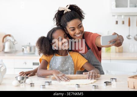 Madre e figlia nere in grembiuli che fanno selfie facendo pasticceria insieme usando il mattarello mentre sono in piedi a tavola con diverse forme di biscotti ... Foto Stock