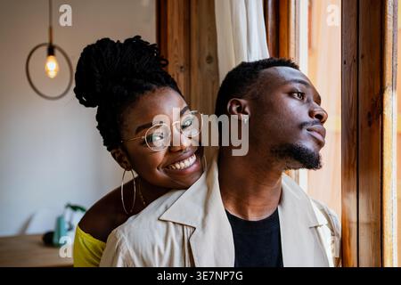Donna afroamericana sorridente negli occhiali che appoggiava il mento all'uomo vicino alla finestra Foto Stock