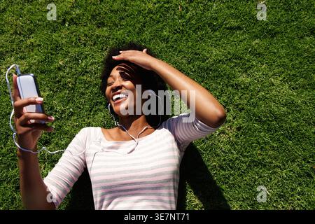 Allegra donna nera con acconciatura afro che indossa gli auricolari e utilizza lo smartphone steso in erba con le mani che coprono il viso dalla luce del sole Foto Stock