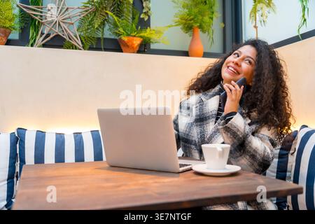 Donna ispanica sorridente con capelli ricci che fa una telefonata seduta al tavolo con un computer portatile in un bar Foto Stock