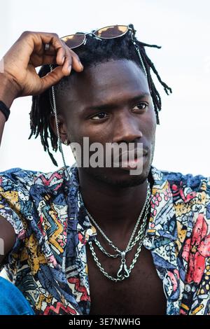 Uomo nero serio che indossa un'elegante camicia, una collana e occhiali da sole sulla testa, con lo sguardo lontano seduto su sfondo bianco in studio Foto Stock