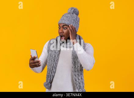 Scioccato Black man che indossa berretto e sciarpa in maglia guardando lo smartphone leggendo notizie sconvolgenti su sfondo giallo in studio Foto Stock