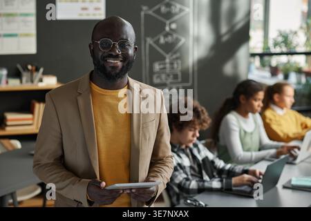 Istruttore di uomo Nero sorridente che tiene un tablet in piedi contro diversi gruppi di studenti nel laboratorio di computer durante il giorno Foto Stock