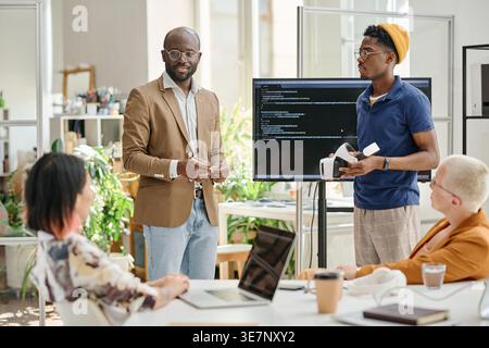 Programmatori maschi neri che testano nuovi occhiali per realtà virtuale in piedi vicino al monitor con codici che tengono una riunione con colleghi in ufficio Foto Stock