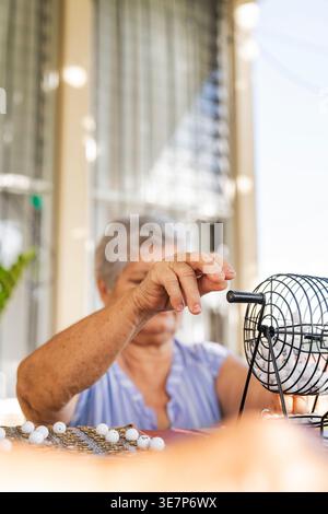 Donna anziana ispanica che mescola le palle nella culla del bingo mentre gioca a casa nelle giornate di sole Foto Stock