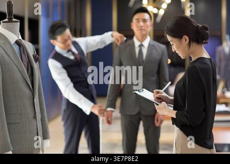 Stilisti cinesi maschili e femminili che misurano i clienti cinesi maschili mentre si trovano in una boutique di moda Foto Stock