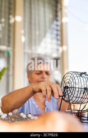 Donna anziana ispanica che mescola le palle nella culla del bingo mentre gioca a casa nelle giornate di sole Foto Stock