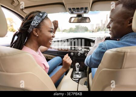 Allegra donna nera che si gode un viaggio in auto con il fidanzato che la guarda mentre guida l'auto durante il giorno Foto Stock