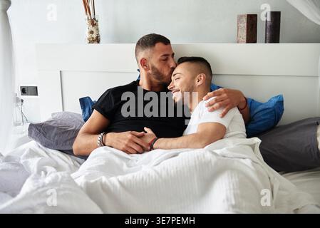 Coppia gay ispanica e caucasica che si coccola sdraiata sul letto a casa Foto Stock