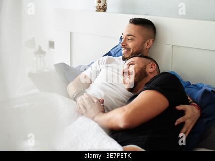 Coppia gay ispanica e caucasica che si coccola sdraiata sul letto a casa Foto Stock
