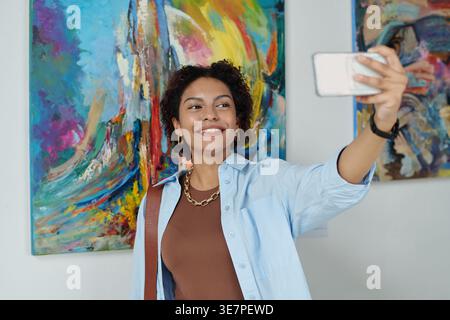 Adolescente nera che scatta selfie contro i dipinti mentre è in piedi nella galleria d'arte Foto Stock