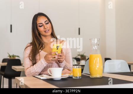 Donna ispanica sorridente che usa lo smartphone mentre siede a tavola e fa colazione in hotel Foto Stock