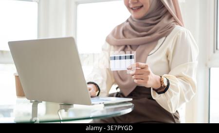 Donna musulmana sorridente che indossa hijab usando la sua carta di credito per acquistare online usando il laptop mentre si siede a un tavolo di vetro vicino alla finestra di casa Foto Stock